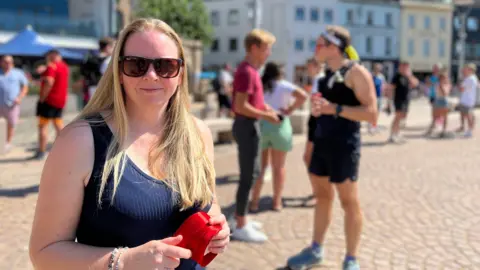 Niamh has long hair stands in a sunny public square holding a red object. Behind her, people walk and talk near buildings under a clear blue sky
