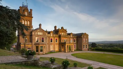 Chris Lacey Photography The front aspect of Hestercombe House, a grand, beige-gold stoned building that has a tower to the left. In front of the house is a large driveway with an oval grassy lawn. Fields and trees extend into the distance.