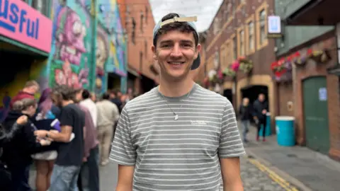 BBC A man stands smiling on a street. He wears a baseball cap backwards and a grey and white striped shirt. There is a queue behind him.
