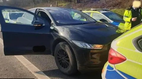 North West Motorway Police A black Jaguar I-Pace car parked on the motorway tightly boxed it at the front and rear by marked police patrol cars. An officer in a high visibility coat, with his face blurred, is standing on the passenger side of the vehicle.