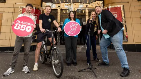 Glasgow City Council Five individuals posing in front of the Glasgow Film Theatre. They are holding signs. One sign says "Glasgow 850" in large numbers on a red circle. Another sign features a pink circle with a banana illustration containing what appear to be the letters "BC". There's a bicycle in the photo too.