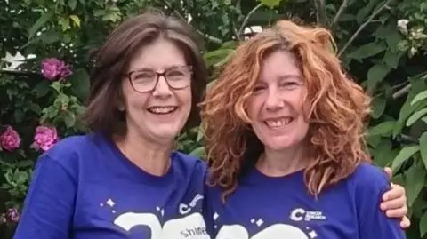 Cancer Research UK Jill and Jan smile as they embrace with their arms around each other for the camera. They both wear blue Cancer Relief UK tops as they stand in a garden with pink roses and green shrubs behind them. Jill has dark shoulder-length hair and wears glasses. Jan has wavy ginger hair.
