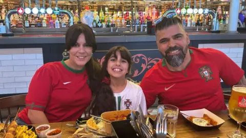 Family photograph Alexandra, Alice and Sergio Aguiar in a restaurant eating a meal al wearing Portugal football shirts. They are all smiling.