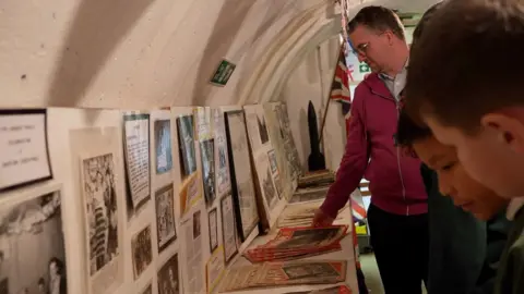 Students looking at World War Two artefacts inside the Clifford Road tunnels. Pictures and documents rest on shelves and against a wall. 