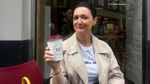Mimoza Emsa with dark brown hair in a ponytail wearing a beige jacket over a white t shirt sitting outside Pret holding a takeaway coffee cup