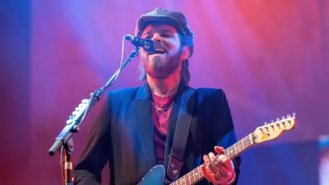 BBC Gaz Coombes on stage at Belladrum. He has a beard and shoulder length hair. He is wearing a cap and a dark suit jacket over a red t-shirt. He has an electric guitar slung from his shoulders as he sings into a microphone.