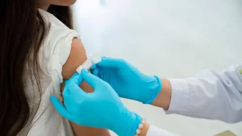 Getty Images A female child with long brown hair and a white top is getting a white plater stuck onto her arm following an injection. They person putting the plaster on is wearing light blue gloves, a pale blue top and has a pearl bracelet on one of their wrists. 