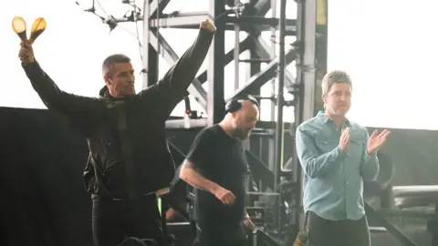 Getty Images Two men raise each others hands. The man on the left is wearing a dark green raincoat and holds two shakers in his right hand. The man on the right wears a blue denim shirt, top button un-done, They both have short brown hair. Behind them are several men who stand in front of a screen displaying green patterns.