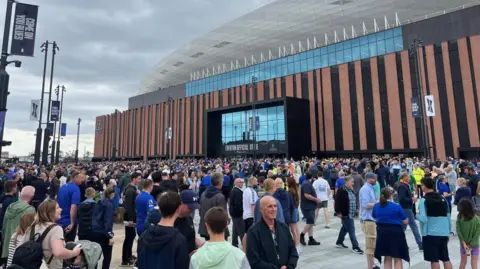 BBC Hundreds of fans, many wearing Everton colours stand in the plaza outside the Hill Dickinson stadium, ahead of the match with Roma. The facade of the stadium features stripes of red brick and black windows and is dominated by a plate-glass entrance.