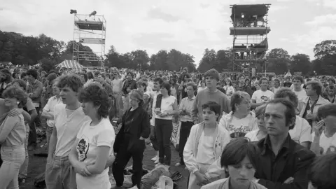 Mark Hodgson A black-and-white shot of a large crowd of people, mostly young in their teens and 20s, with 1980s clothing and hairstyles. It is an outdoor concert in daylight, and they are all looking towards the stage which is out of shot to the left of frame. Behind them in the background are two scaffolding towers, and a line of trees.