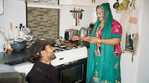 Jay Singh Jay Singh is sat in his grandma's kitchen, head height with the kitchen counter. He is wearing a black cap and a black turtleneck jumper. He is looking up at his grandma as she cooks on the hob while wearing green and pink traditional south Asian clothing. In the kitchen are various common kitchen item such as a kettle, toaster, oven and kitchen knives.