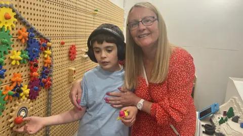 A woman with blonde hair wearing a red spotty dress standing with her son who has headphones on