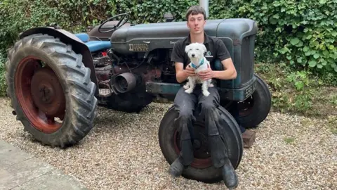 Billy Kitcher Billy Kitcher sat on his tractor's front tyre holding his small white dog.