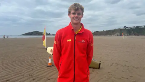 A young male lifeguard wearing a red fleece standing on a sandy beach in front of an RNLI flag. The sea is in the distance behind him.
