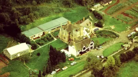 Other An aerial picture of Berrow Wood School with fields surrounding a black and white Tudor manor. There are cars parked on the road outside and bushes and trees around the edge