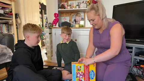 Roxanne and her two boys sitting on the floor as she opens a new game 