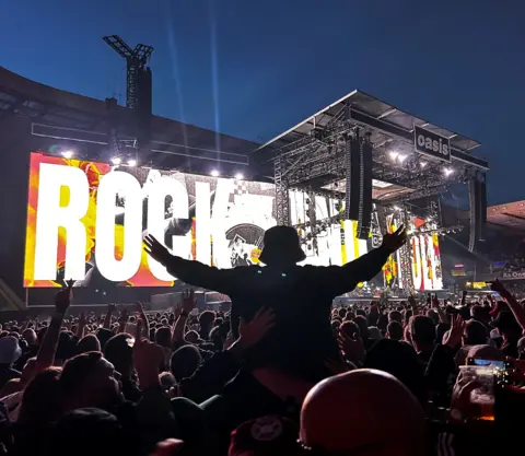 E Lamb Large crowd at a concert photographed from behind - one person is raised in the air with their arms out, wearing a bucket hat with a large display on stage screens reading "Rock N Roll"
