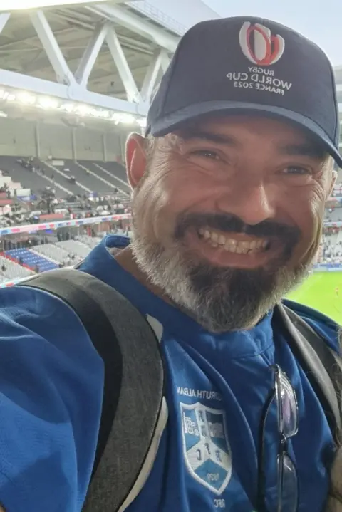 Simon Ferguson A selfie of Simon Ferguson as he is in stands overlooking a rugby pitch. He is wearing a blue cap with a logo and WORLD CUP printed on it and a blue club shirt. Grey backpack straps are visible across his shoulders. He is grinning and has a dark and grey beard and brown eyes.