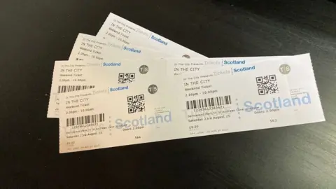 Four white In The City festival tickets on a black wooden surface. The tickets have black text, barcodes and blue logos on them.