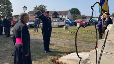 Men in suits and military uniforms gather sparsely near an engraved stone war memorial that can just be seen to the right. A man is saluting while facing the memorial. Men can be seen holding colourful flags.