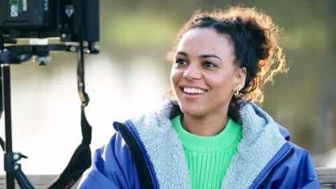 Rob Youngson Photography Cat White, a filmmaker, sitting down looking into a camera monitor. She is wearing a bright green jumper with a blue fleece-lined coat over the top. She is smiling but not looking at the camera.