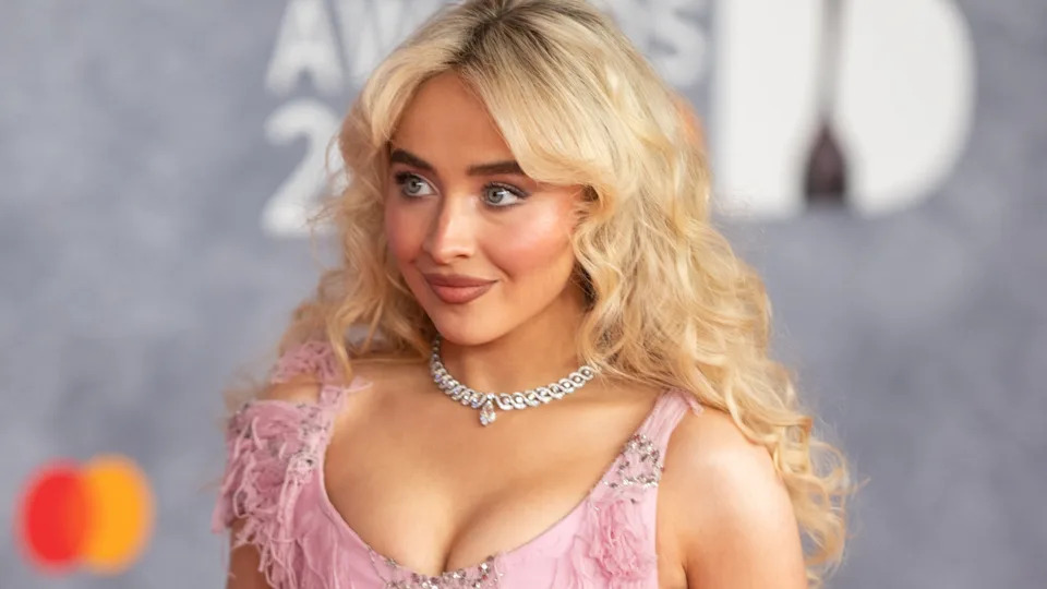 Sabrina Carpenter with long blonde hair in a pink dress and necklace, posing at an event with a blurred background.
