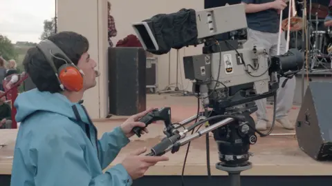 Mark Hodgson A colour mid-shot side view of a camera operator and a television camera, outside in daylight just in front of a large stage. The cameraman appears to be approximately in his 30s, has dark hair, and is seen left of frame wearing red headphones and a pale blue rain jacket. He is holding the operating handles of his large television camera and looking into its viewfinder. The camera is fixed onto a mounting and has an Anglia Television logo on its side.