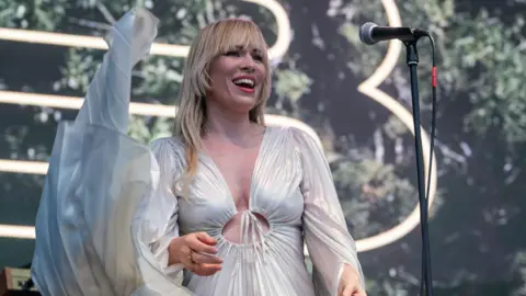 BBC ALBA Natasha Bedingfield is pictured performing on Belladrum's main stage. She has long blonde hair and is wearing a white, flowing dress. She is smiling as she sings into the microphone.