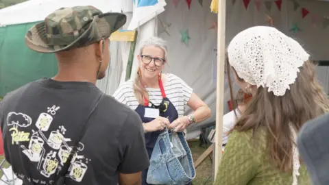BBC Clare seeking fixing a denim skirt in front of two festival attendees. She is wearing an apron and striped t-shirt.