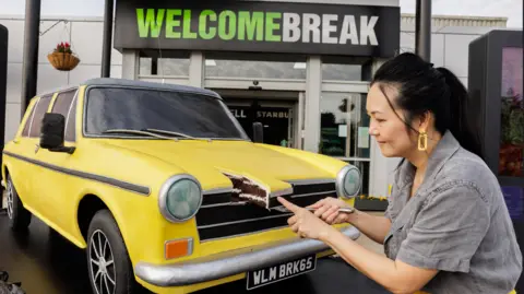 Welcome Break A life-like car made of cake outside a Welcome Break service station. The car's design is based on an Austin/Morris from 1960 and is yellow in colour. A woman with black hair and wearing a grey top is cutting a slice out of the bonnet.