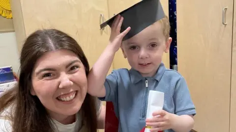 PA Media Conor holds his little black graduation mortar board hat on his head. Cupboards are behind him in a nursery.