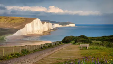 BBC Picturesque white cliffs, known as the Seven Sisters, face a pale blue ocean. The sun is setting.