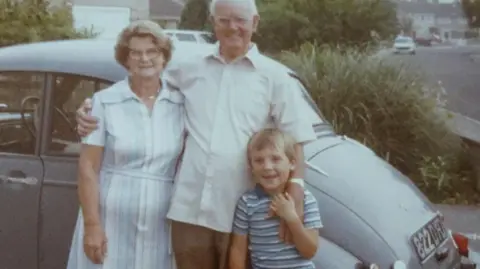 BBC An old photograph of an elderly couple standing in front of a grey classic car with their grandson. The man has his arms around the woman and the child. All of them are smiling. 