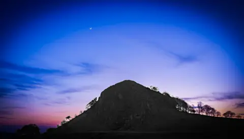 Bartek Kaluzny Landscape image of the shadow of a hill and trees leading down with the vivid background of a blue, purple, and pink ombre sky and crescent moon