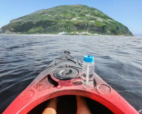John Dewar Red kayak with a water bottle on the rim, floating in the water from the point of view of the kayaker with the Ailsa Craig in the distance