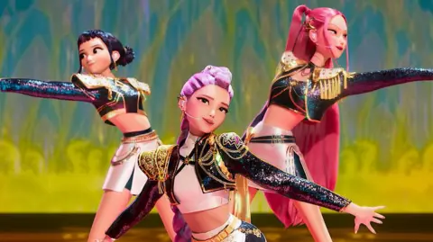 Netflix A scene from the animation K-pop Demon Hunters, where three young women - kpop idols - are dressed in shiny costumes and performing on a stage. They are posing with their arms outstretched.
