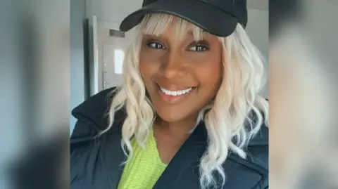 Rhakima Kahn A woman is seen close up. She is smiling at the camera. She is wearing a green jumper under a black jacket and a black cap.