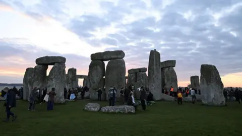 Reuters People gathered around the Stonehenge as the sky turns a pinky orange. There is a light layer of cloud. 