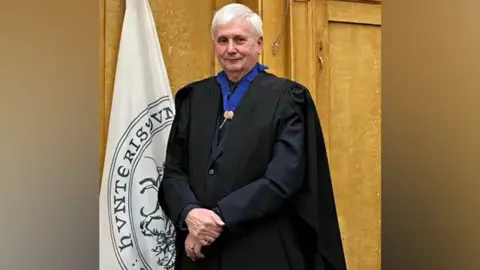 Huntingdon Town Council Steve McAdam is wearing a dark blue collar, a black jacket and a long blank gown. He is standing in front of a wooden doorway and white flag with blue lettering. 