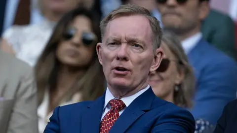 Getty Images A man with short grey-brown hair, wearing a blue suit, white shirt and red tie, sat with a face of concentration in the stands at Wimbledon.