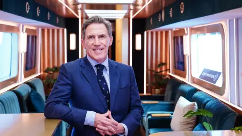 PA Media Rob Brydon standing in a blue suit in a carriage with blue seats behind him.