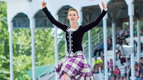 Kathy Park photography The image shows a woman performing a Highland dance. She is mid-leap, arms outstretched. She is wearing a traditional Highland dance costume: a dark velvet jacket with white accents, a pleated tartan kilt in shades of purple and white, and matching patterned socks and shoes.