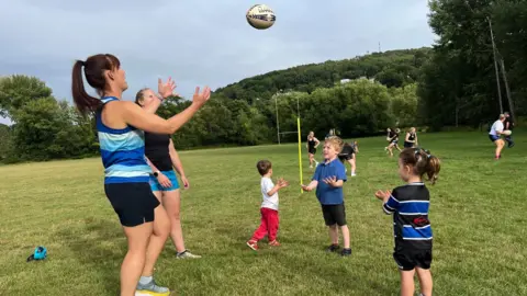 Two women, one about to catch a rugby ball, playing with three young children. One is in a rugby kit, the others are in casual outfits. They are standing on a rugby pitch with people training in the background.