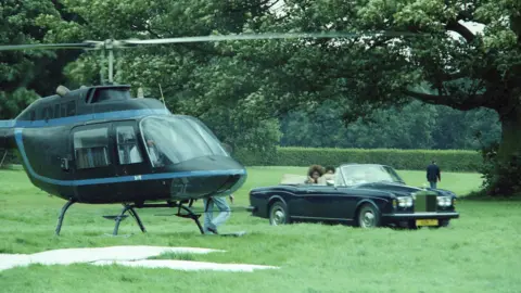 Mark Hodgson A colour shot of a green field with trees in the background in daylight, with to the left of frame a black helicopter with its rotors moving having either just landed or preparing for take-off. To the right of frame is an open-topped Rolls-Royce, with three women being driven in it as passengers. Next to the helicopter slightly to the foreground appears to be a large white 'x' shape marked out with sheets or boards.