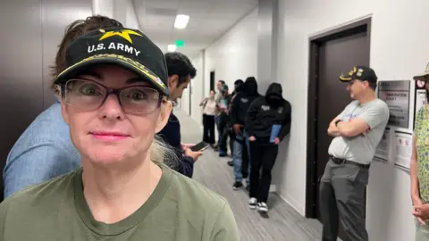 BBC Monique stares at the camera, wearing a black baseball cap that says US Army, and an olive-green t-shirt. In the background is a fluorescent hallway and a man in a gret t-shirt and the same black army cap. Several ICE officers, masked and in black hoodies, are also in the hallway