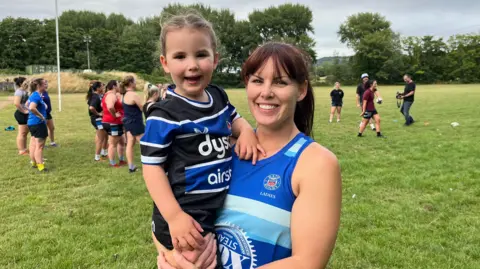 Samantha Castley-MacDonald in a blue Bath Ladies vest, holding her daughter who is in a small blue and black rugby kit. They are both smiling and there are people training behind them.