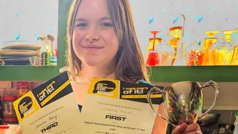 Hazel Davis A 13-year-old girl holds two certificates and a trophy and smiles at the camera