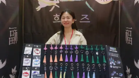 Choi Nyun-hee Choi Nyun-hee smiles at the camera, wearing a yellow collared shirt. She has long black hair and a red lanyard. Perched in front of her is a black board with keychains and bracelets hanging on it.