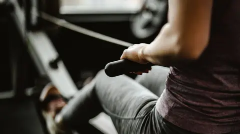 Getty Images A woman in gym clothing is seen on an indoor rowing machine. 
She is wearing a light purple top and grey sport leggings. 
