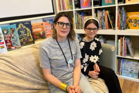 A woman and a young female child sat on a sofa. The woman is on the left and has shoulder-length brown hair and round glasses. She is holding her hands in her lap, and is wearing a grey T-shirt, faded blue jeans, and a gold strap around her right arm. The girl is on the right and is wearing similar round glasses and a white headband, and has brown hair tied in a ponytail. She is wearing a baggy black jumper with white flowers on it, and black trousers. They are sat on a grey sofa covered in a cream-coloured blanket. There are children's books on display behind the sofa, and on the right hand side there is a set of book shelves filled with colourful children's books.